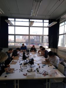 A room with 8 people working at a table covered in wires