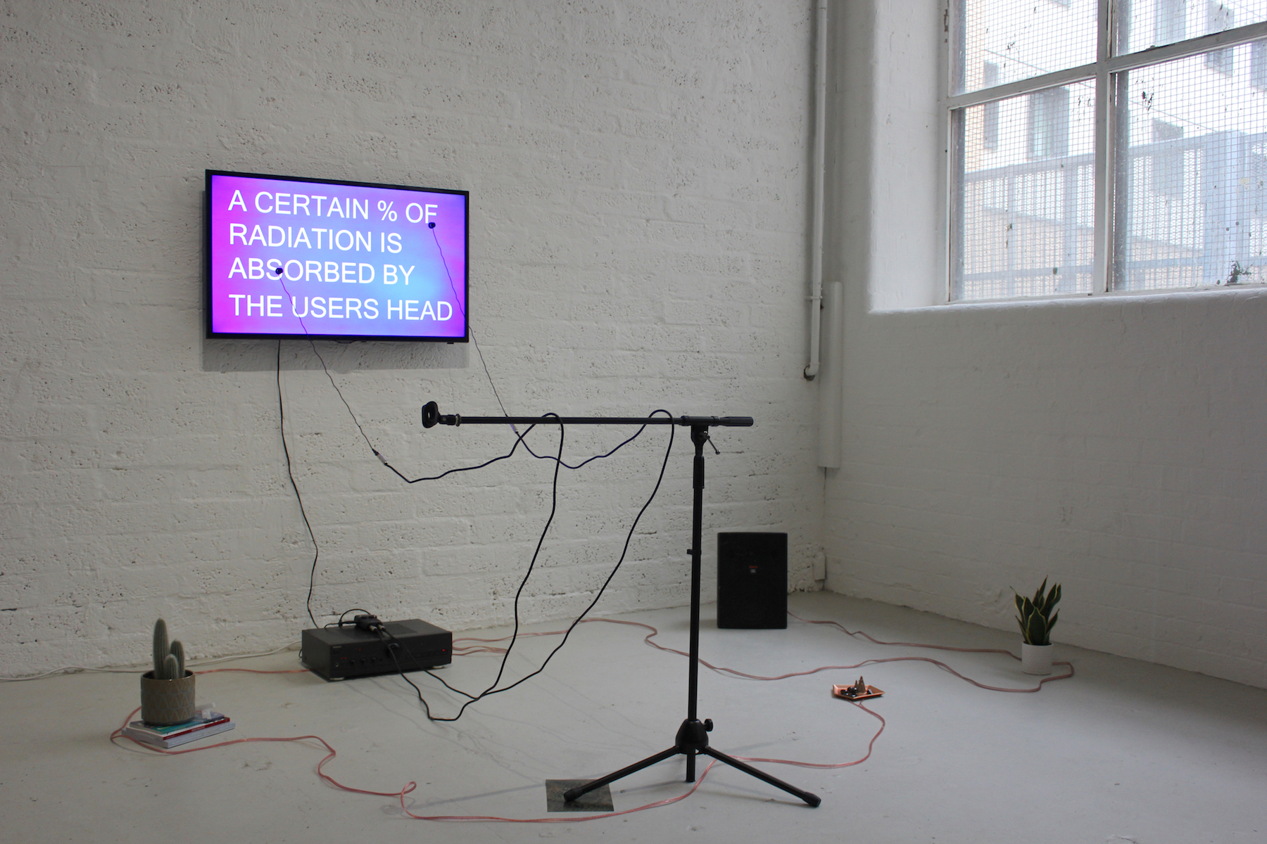 a microphone stand with cables and a tv screen on the wall with white text on pink, reading " A CERTAIN % OF RADIATION IS ABSORBED BY THE USERS HEAD"