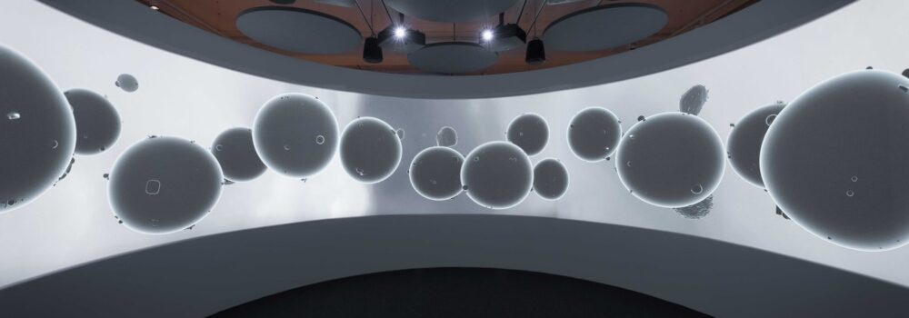 panoramic film installation in a round room with speakers overhead - the screen shows a wide white surface with numerous round objects